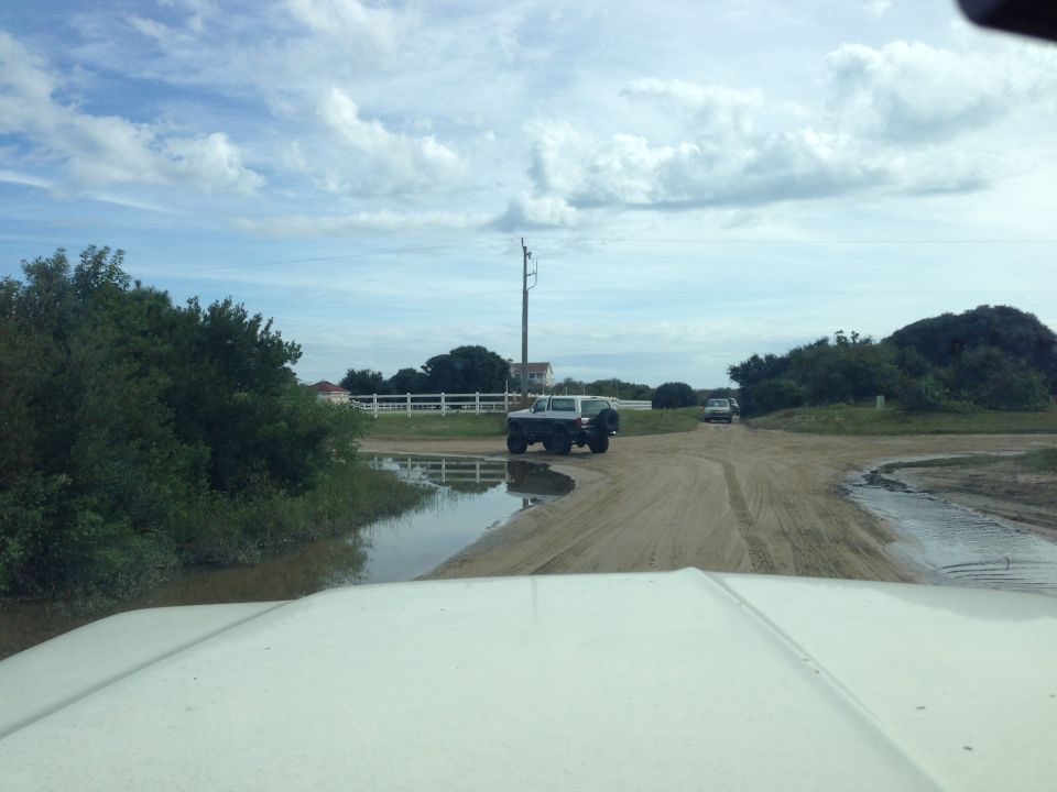 White SUV on a dirt road with a puddle, viewed from the front.
