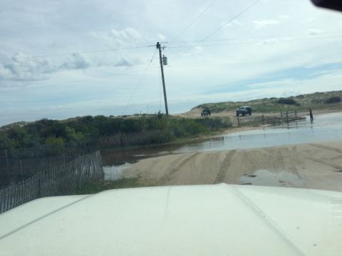1979 Ford Bronco - The front of a white vehicle is visible in the foreground, looking out at a sandy road with puddles. Two SUVs are driving on the road ahead…