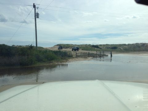 1979 Ford Bronco - The front of a white vehicle is visible, driving through a shallow body of water on a sandy path. Two dark-colored SUVs are visible further…