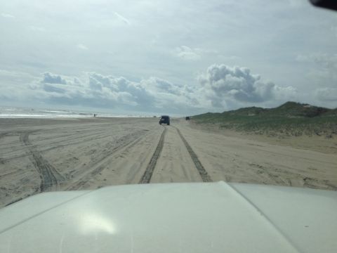 1979 Ford Bronco - The front of a white vehicle is visible in the foreground, looking out at a sandy beach with tire tracks. Two dark SUVs are driving on the…