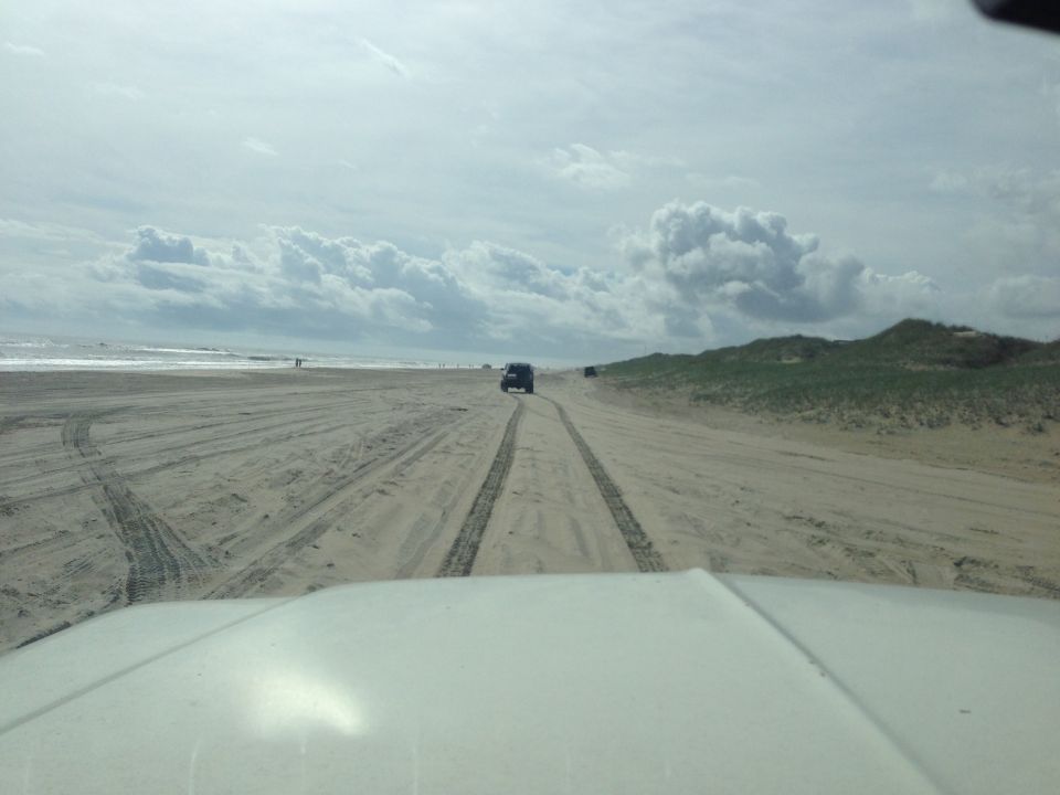 White vehicle hood in foreground, looking out at a beach with tire tracks and two distant SUVs.