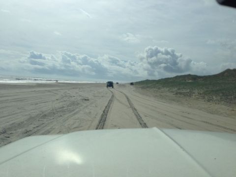 1979 Ford Bronco - The front of a white vehicle is visible, looking out over a sandy beach with tire tracks. Two dark-colored SUVs are driving on the beach in…