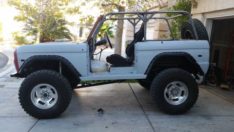 1979 Ford Bronco - A light gray, modified Ford Bronco is parked in a driveway with a roll cage and large off-road tires. The vehicle is missing its doors and…