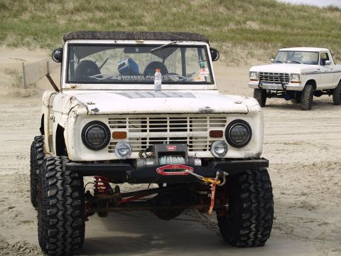 1979 Ford Bronco - A weathered white Ford Bronco with large off-road tires and a winch is parked on a sandy beach. A white Ford pickup truck is parked behind…