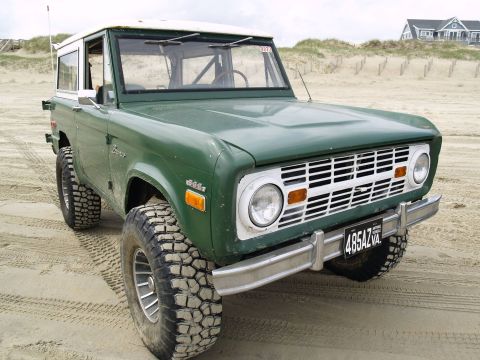 1979 Ford Bronco - A green 1970s Ford Bronco with large off-road tires is parked on a sandy beach, with dunes and a house visible in the background.