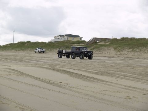 1979 Ford Bronco - A black Ford Bronco with a trailer is driving on a sandy beach, with a white pickup truck in the distance. A yellow house and wooden stairs…