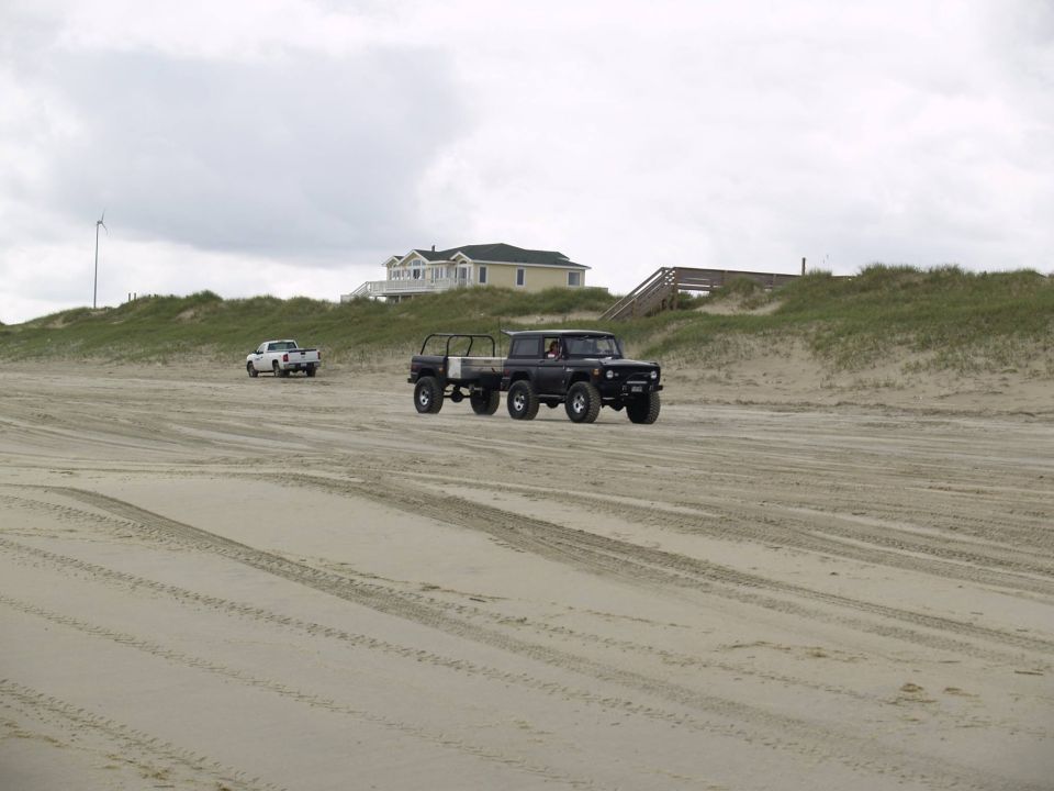 Black Ford Bronco with trailer driving on a sandy beach, white pickup in distance.