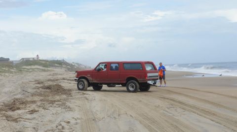 1979 Ford Bronco - A red, four-door SUV is parked on a sandy beach near the ocean. A man in a blue shirt and shorts stands next to the rear of the vehicle hol…