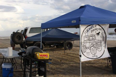 1979 Ford Bronco - A white Ford Bronco is parked on a sandy beach under a blue canopy, with a banner displaying "TIDEWATER BRONCOS" and a drawing of a classic…