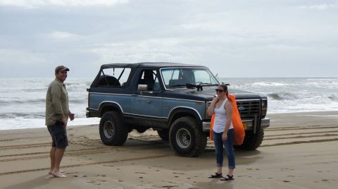 1979 Ford Bronco - A man and a woman stand on a sandy beach next to a blue and black Ford Bronco with a soft top. The ocean waves are visible behind them.