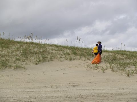 1979 Ford Bronco - Two people are walking up a sandy dune covered in sea grass under a cloudy sky. One person is holding an orange bag.