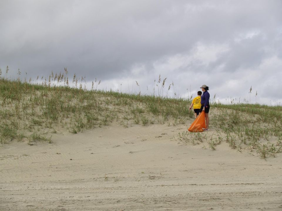 Two people walking up a sandy dune with sea grass under a cloudy sky.