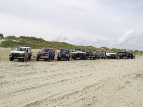 1979 Ford Bronco - A line of vintage Ford Broncos and trucks are parked on a sandy beach under a cloudy sky. Several people stand around the vehicles.