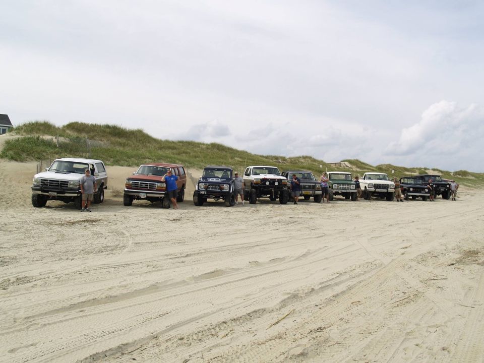 A line of vintage Ford Broncos and trucks parked on a sandy beach.
