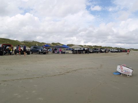 1979 Ford Bronco - A group of people and several vehicles are gathered on a sandy beach with dunes and houses in the background. A red off-road vehicle is par…