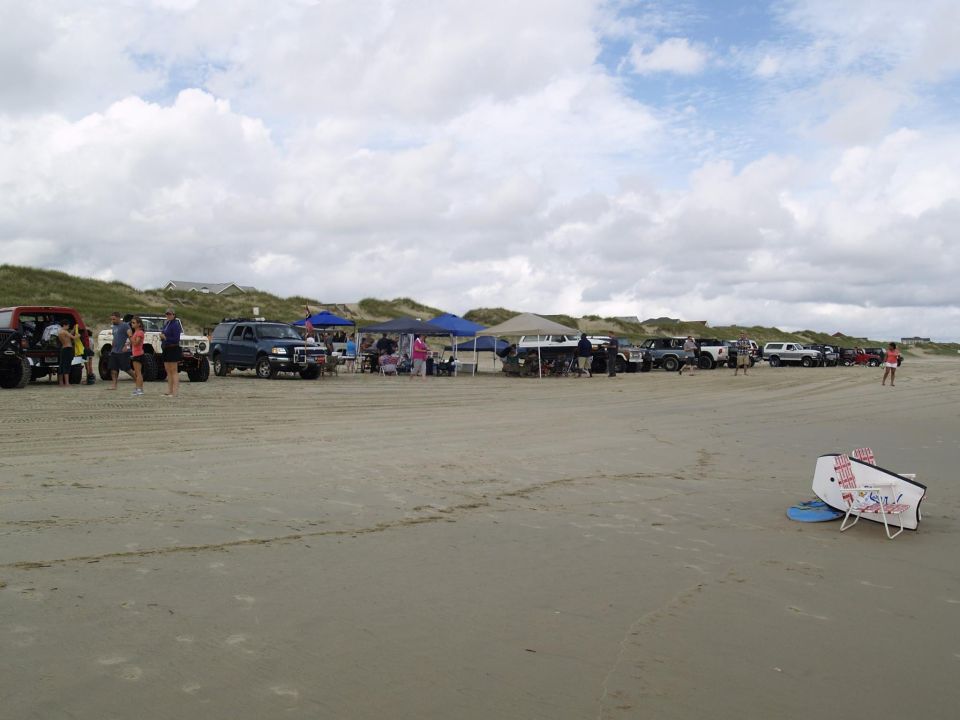 Red off-road vehicle and other trucks on a sandy beach with people and dunes in the background.