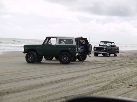 1979 Ford Bronco - Two Ford Broncos are parked on a sandy beach with the ocean in the background. The Bronco in the foreground is dark green with a white top,…