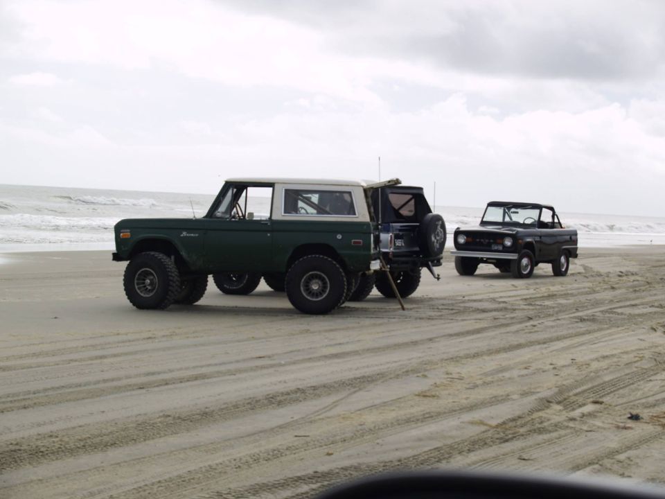 Two Ford Broncos on a beach, one green and white, one black.
