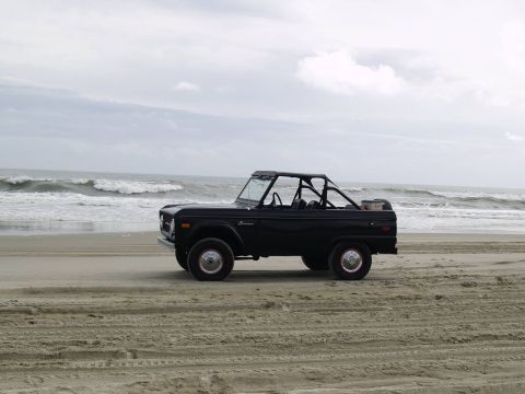 1979 Ford Bronco - A black, convertible 1979 Ford Bronco sits on a sandy beach facing the ocean. The vehicle has a roll cage and red-accented wheels.