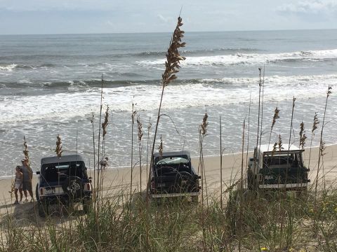 1979 Ford Bronco - Three off-road vehicles are parked on a sandy beach near the ocean. Two black Jeeps are visible in the foreground, with a green Jeep visibl…