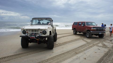 1979 Ford Bronco - A white 1979 Ford Bronco and a red Ford F-Series SUV are parked on a sandy beach near the ocean under a cloudy sky. The Bronco has a winch…