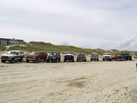 1979 Ford Bronco - A line of SUVs and trucks are parked on a sandy beach with dunes and a house in the background.