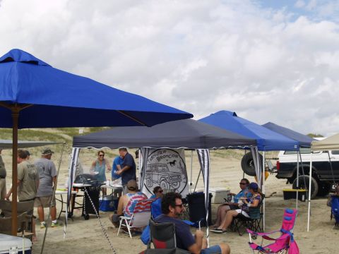 1979 Ford Bronco - A group of people are gathered on a sandy beach under several canopies, with a dark blue and white Ford Bronco visible in the background. T…