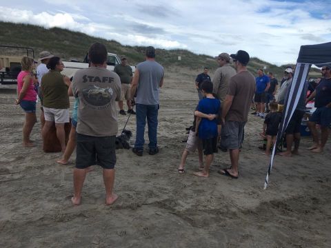 1979 Ford Bronco - A group of people are gathered on a sandy beach with dunes in the background. A white truck is partially visible behind some of the people.