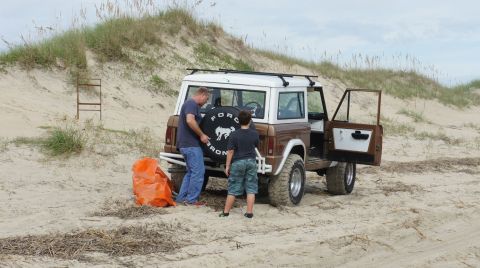1979 Ford Bronco - A man and a boy stand next to a brown and white Ford Bronco on a sandy beach with dunes in the background. The spare tire cover is visible,…