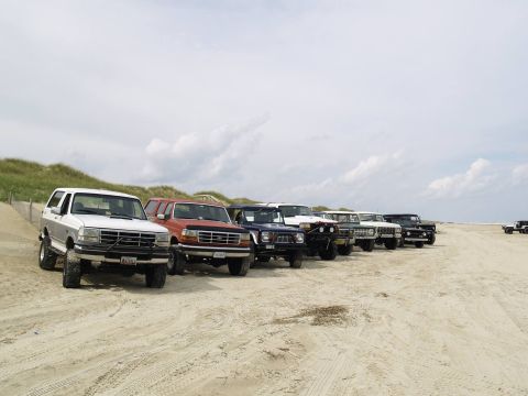 1979 Ford Bronco - A line of SUVs and trucks are parked on a sandy beach with dunes and vegetation in the background under a cloudy sky.