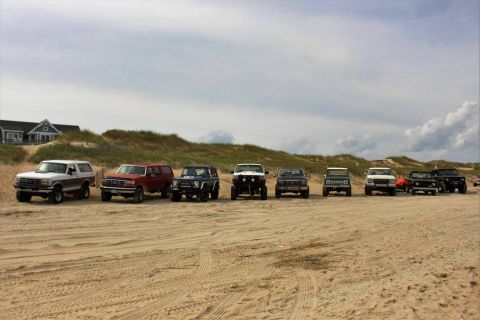 1979 Ford Bronco - A line of SUVs and trucks are parked on a sandy beach with dunes and a house in the background under a cloudy sky.