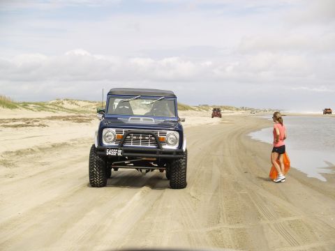 1979 Ford Bronco - A blue Ford Bronco is parked on a sandy beach, with a woman standing nearby holding an orange bag. Other vehicles are visible in the distan…
