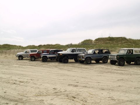 1979 Ford Bronco - A line of six classic Ford Broncos are parked on a sandy beach with dunes and a wooden fence in the background. The vehicles range in color…