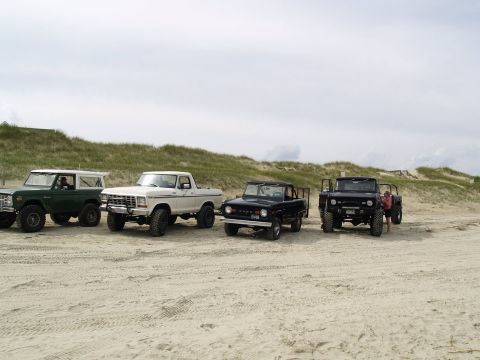 1979 Ford Bronco - Four off-road vehicles are parked on a sandy beach with dunes and vegetation in the background. From left to right, a green SUV, a white pi…