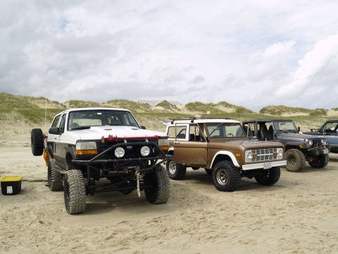 1979 Ford Bronco - A line of four-wheel-drive vehicles, including a white Ford Bronco and a brown Ford Bronco, are parked on a sandy beach with dunes and a cl…