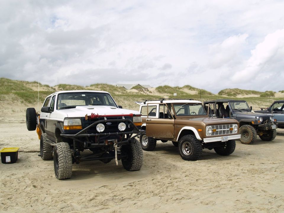 Four off-road vehicles parked on a sandy beach with dunes in the background.