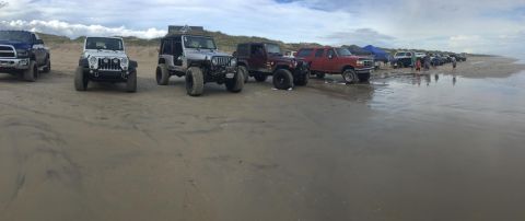 1979 Ford Bronco - A line of off-road vehicles, including Jeeps and trucks, are parked on a sandy beach with dunes and the ocean in the background.