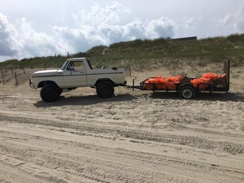 1979 Ford Bronco - A white 1970s Ford Bronco is parked on a sandy beach, towing a trailer loaded with orange bags.