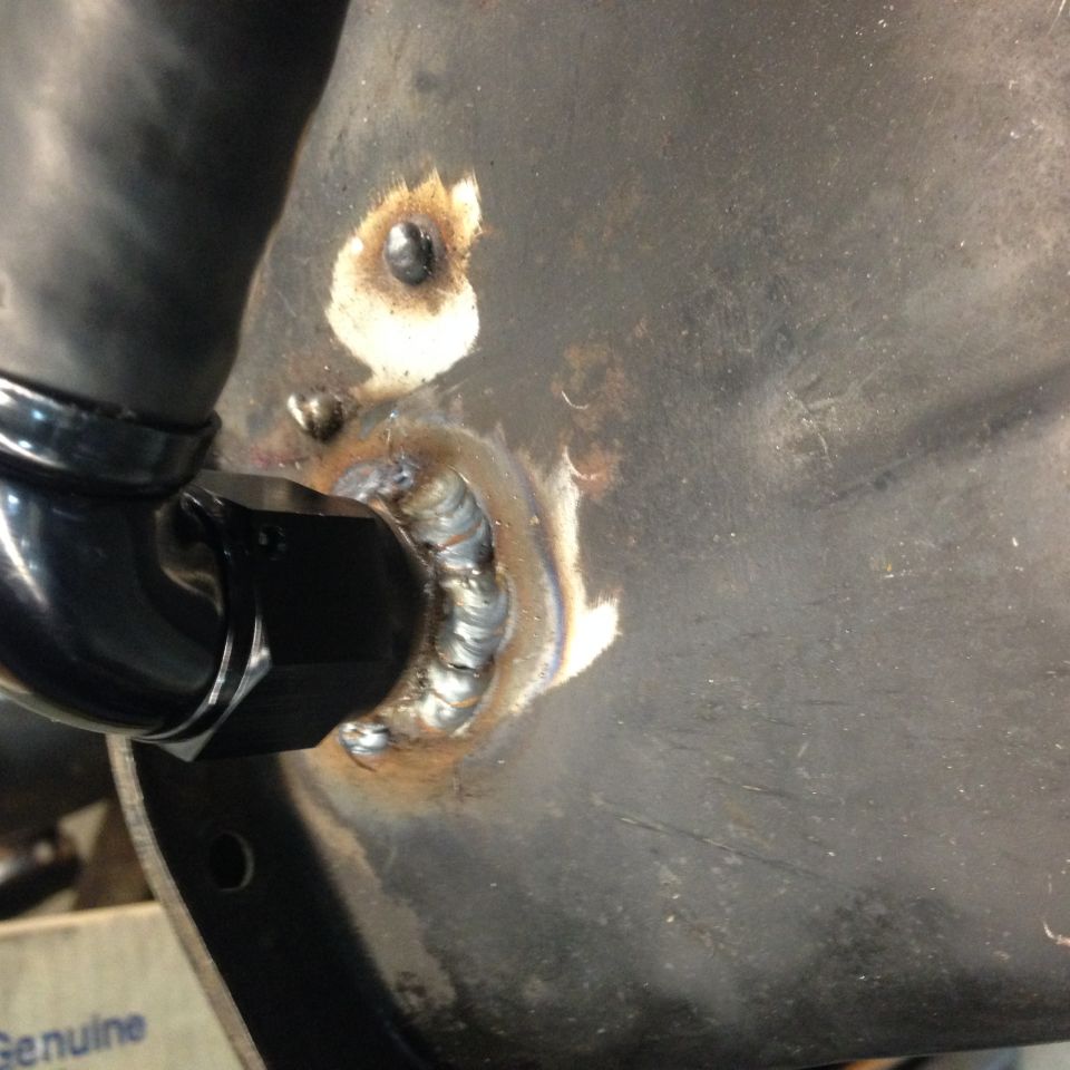 Close-up of a welded fitting with a black hose attached to a dark metal surface.