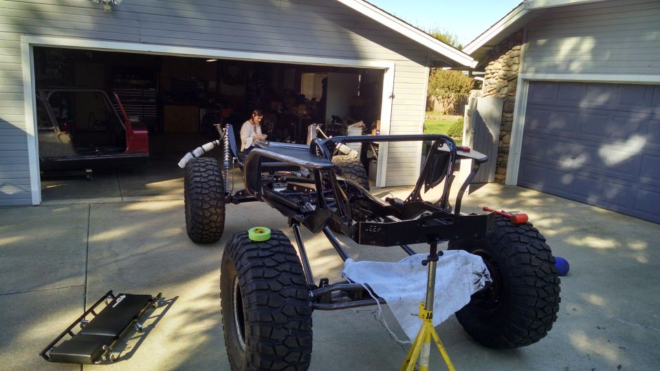 Custom off-road vehicle frame with large tires and a person working inside, parked in front of a garage.