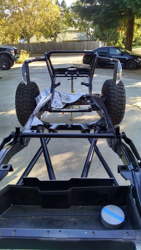 1985 Jeep CJ7 - The bare frame of a custom off-road vehicle, likely a Jeep CJ7, is shown from a low-angle rear view. Large off-road tires are mounted, and…