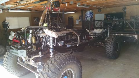 1985 Jeep CJ7 - A custom-built Jeep chassis with a large engine suspended above it by a hoist in a garage setting. The front of the Jeep features large tir…