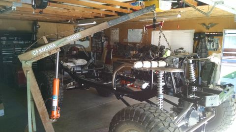 1985 Jeep CJ7 - A Jeep CJ7 frame is being worked on in a garage. An engine is suspended by a shop crane, ready to be installed into the frame.
