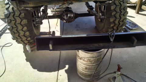 1985 Jeep CJ7 - The rear axle and suspension of a Jeep CJ7 are visible, with a custom steel bumper being prepared for installation. A large tire and part o…