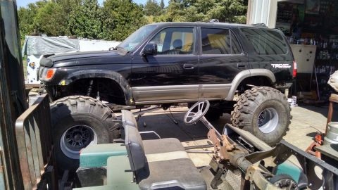 1985 Jeep CJ7 - A modified black Toyota 4Runner SUV with oversized tires and a lifted suspension is parked outdoors next to a trailer and a workshop. A ste…