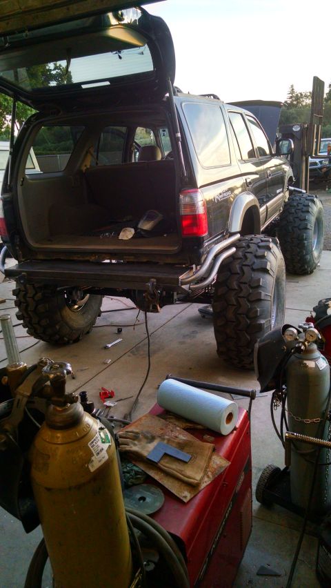 1985 Jeep CJ7 - A heavily modified black SUV with oversized tires is parked in a garage or workshop. Welding equipment and tools are scattered around the v…