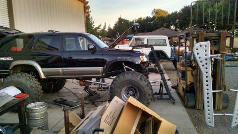 1985 Jeep CJ7 - A black Toyota 4Runner with oversized tires and lifted suspension is parked outside a garage next to a white Jeep SUV. A forklift and vario…