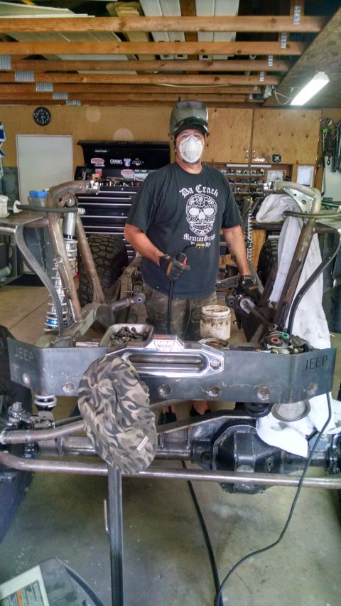1985 Jeep CJ7 - A person wearing a welding mask and protective gear stands in a garage, working on the front suspension and bumper of a Jeep CJ7. The vehic…