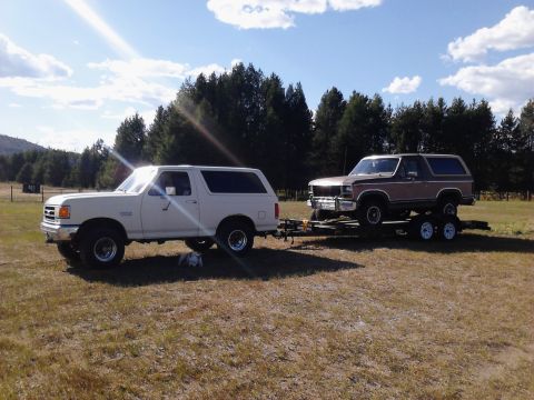 1990 Ford Bronco - A white Ford Bronco is parked in a grassy field, towing a trailer carrying a brown and tan Ford Bronco. The sky is blue with scattered clou…