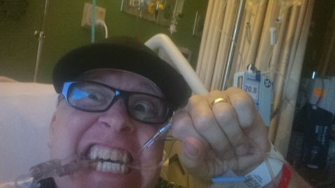 1985 Jeep CJ7 - A person wearing glasses and a hat is in a hospital bed, holding a nasal cannula to their mouth. Medical equipment, including an IV pump, i…